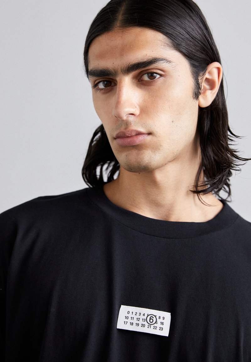 T-shirt in cotone nero con collo rotondo, caratterizzato da un'etichetta bianca sul petto che mostra numeri e un cerchio, con il numero 6 evidenziato.