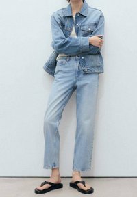 Chaqueta de denim azul claro y jeans de corte recto, llevados sobre una blusa crema, combinados con chanclas negras sobre un fondo neutro.