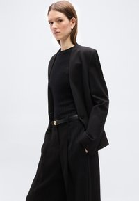 Νεαρή γυναίκα που φορεί μαύρο σακάκι, μαύρο ριπ τοπ και μαύρο ψηλόμεσο παντελόνι με λεπτές ρίγες και μαύρη ζώνη, στέκεται με τα χέρια στις τσέπες.