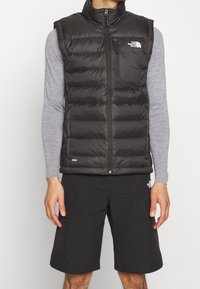 Muž v černé péřové vestě, šedém dlouhém rukávu a černých šortkách, vše značky The North Face.