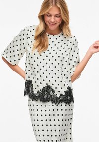 Blouse blanche à pois noirs, avec manches courtes et ourlet en dentelle noire. Texture satinée lisse, coupe décontractée.