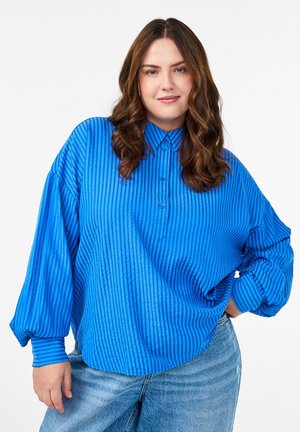 Jonge vrouw met golvend bruin haar, gekleed in een losse blauw gestreepte blouse en lichte blauwe jeans, staat met één hand op haar heup.