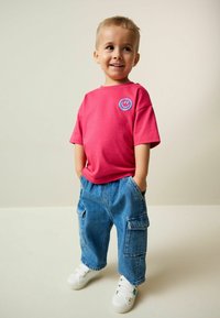 Barn iført en lyserød bomulds T-shirt med blåt smiley-logo, kombineret med løstsiddende blå denim cargobukser og hvide sneakers.