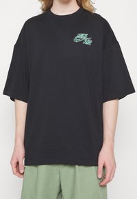 Svart oversized t-shirt i bomull med en grön grafisk logga på bröstet. Kortärmad och avslappnad passform.