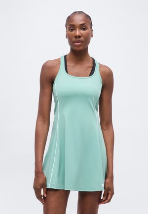 Femeie atletică purtând o rochie verde mentă fără mâneci, cu un sutien sport negru pe dedesubt, stând în picioare pe un fundal deschis uniform.