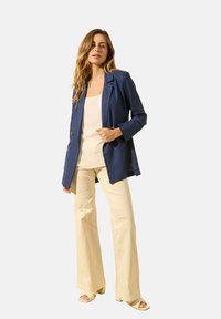 Blazer marine à épaules structurées et boutons, associé à un pantalon beige évasé et un haut clair. Tissu lisse et coupe ajustée.