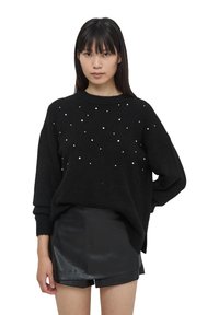 Czarny, dzianinowy sweter z okrągłym dekoltem, ozdobiony rozrzuconymi białymi koralikami, w połączeniu z czarnymi skórzanymi szortami.