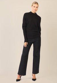 Czarny sweter prążkowany z wysokim dekoltem w zestawieniu z czarnymi spodniami typu wide-leg z delikatnymi srebrnymi akcentami. Modelka nosi czarne szpilki.