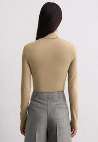 Frau mit langen dunklen Haaren, die ein beigefarbenes langärmliges Oberteil und hoch sitzende graue Hosen trägt, steht mit dem Rücken zur Kamera.