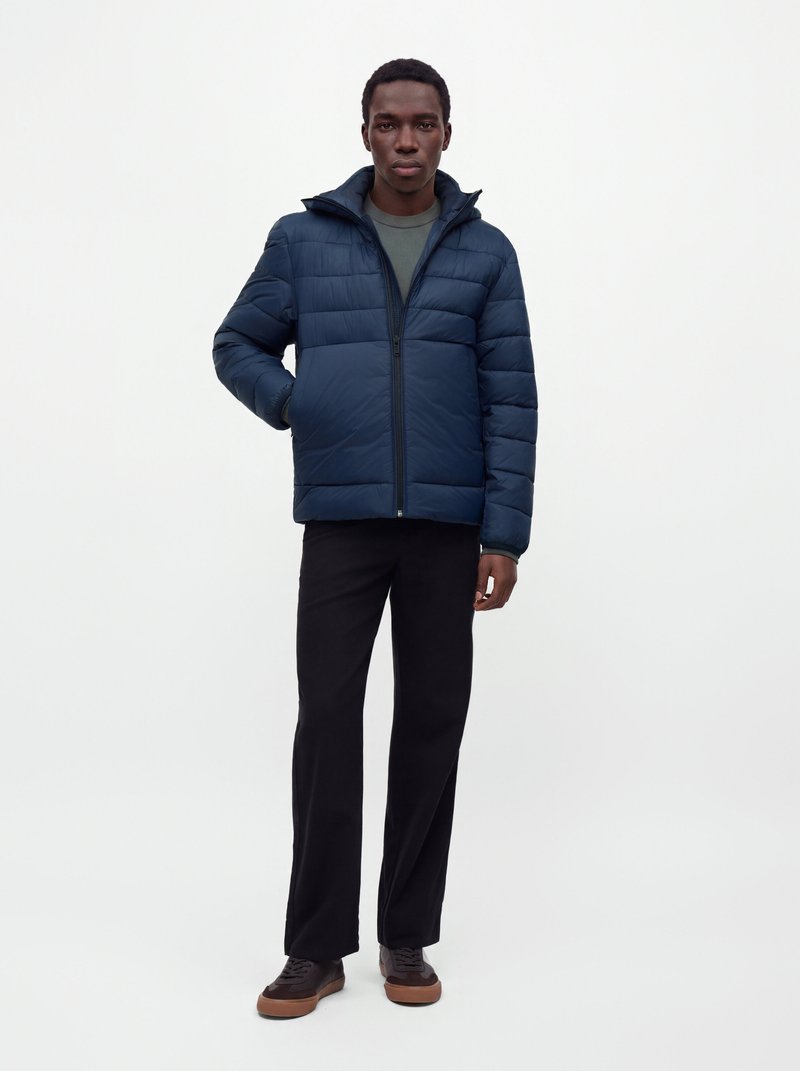 Homme portant une veste matelassée bleu marine, un pantalon foncé et des baskets marron, debout devant un fond blanc uni.