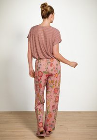 Vrouw met een opgestoken haar, draagt een rood gestreept kortmouwtop en roze broek met bloemenpatroon, staand op een houten vloer tegen een witte muur.