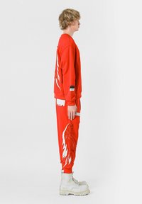 Sudadera roja y pantalones a juego con diseños de plumas blancas; cuentan con un ajuste holgado, puños acanalados y accesorios blancos. Botas blancas gruesas.