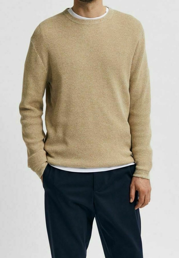 Maglione lavorato a maglia beige con scollo a girocollo, indossato sopra una camicia bianca, abbinato a pantaloni blu navy. Design semplice, senza motivi o loghi visibili.
