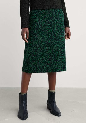 Jupe midi noire avec un motif de feuilles vertes, fabriquée en tissu texturé. Associée à des bottines noires et un pull texturé foncé.