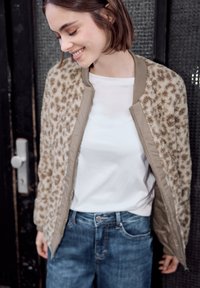 Jeune femme souriante regardant vers le bas, portant une veste imprimé léopard, une chemise blanche et un jean bleu sur un fond sombre et texturé.