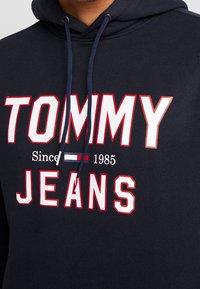 Námořnická mikina s nápisem "TOMMY JEANS Since 1985" v bílé a červené barvě, s malým logem vlajky v červené, bílé a modré barvě umístěným mezi slovy.