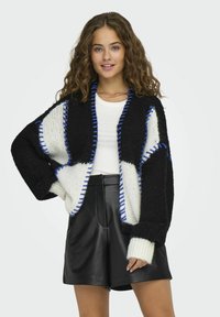 Cardigan de punto en blanco y negro con acentos azules, de corte oversized, de mangas largas y tela texturizada, llevado sobre una blusa blanca y shorts de cuero.
