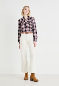 Camicia a scacchi nei colori blu navy, rosso e bianco, con un dettaglio a fiocco; abbinata a pantaloni a vita alta bianchi svasati e stivali platform in suede marrone.
