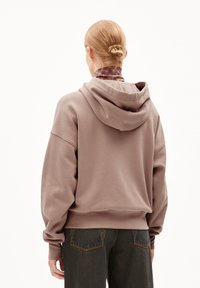 Un sweat à capuche marron à coupe décontractée, avec des épaules tombantes et des poignets côtelés, associé à un jean en denim foncé.