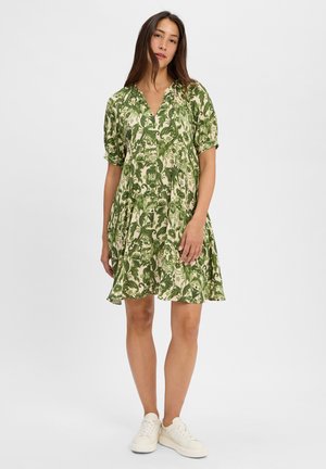 Vestito verde con pattern floreale, scollatura a V, maniche corte e forma ad A, realizzato in tessuto leggero e testurizzato. Indossato con sneakers bianche.