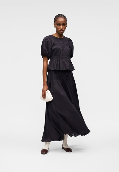 Mannequin portant un haut noir à manches bouffantes et une longue jupe, tenant une petite pochette blanche, avec des chaussettes blanches et des ballerines marron.
