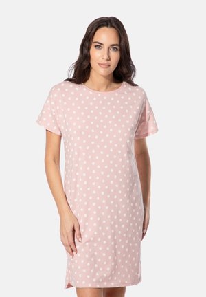 Frau mit dunklen Haaren, die ein pinkfarbenes kurzärmeliges Kleid mit weißen Punkten trägt, steht vor einem schlichten hellen Hintergrund.