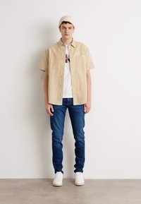 Camicia beige a maniche corte con bottoni sopra una t-shirt grafica bianca, abbinata a jeans blu e sneakers bianche. Il modello indossa un berretto in maglia leggera.