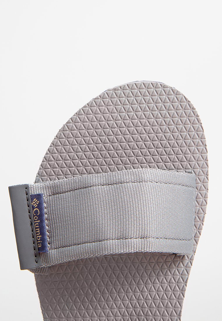 Graue Sandale mit einem strukturierten Gummifußbett mit dreieckigem Muster, ergänzt durch einen grauen Stoffriemen und einem Columbia-Logo-Tag.