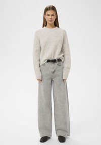 Mladá žena v béžovém chlupatém svetru, širokých džínech s kyselinovým vyblednutím a černým páskem a černých botách, stát před jednoduchým bílým pozadím.