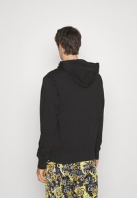 Felpa nera con cappuccio, polsini e orlo a coste, abbinata a pantaloni corti con motivi colorati. Il tessuto sembra morbido e strutturato.