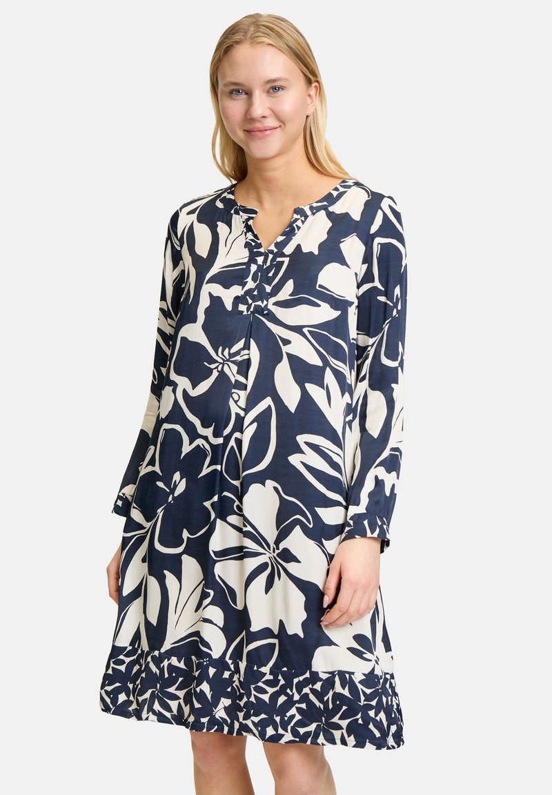 Marineblaues Blumenkleid mit langen Ärmeln, V-Ausschnitt und weißen Blumenmustern. Der Saum hat einen rüschengeschnittenen Rand und besteht aus leichtem Stoff.