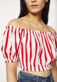 Femme portant un haut court à épaules dénudées à rayures verticales rouges et blanches avec des bandes élastiques aux manches et à la taille, associé à un jean bleu en denim.