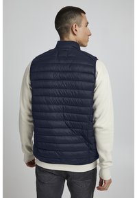 Marineblaues Steppwesten mit horizontalem Steppmuster, hohem Kragen, ärmellosem Design, über einem hellen cremefarbenen Pullover getragen.