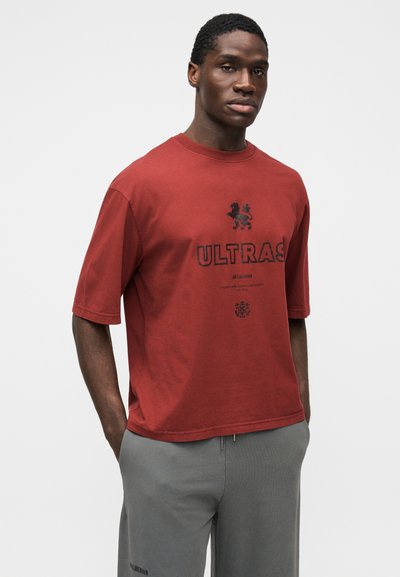 T-shirt en coton bordeaux à manches courtes, avec le texte noir "ULTRAS" et un graphisme de lion. Associé à un pantalon de survêtement gris clair.