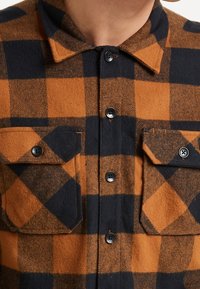 Buffalo geruite shirt met oranje en marineblauwe blokpatroon, wolachtige textuur, knoopsluiting aan de voorkant en twee voorzakken met kleppen.