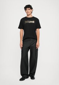 Melnas kokvilnas T-kreklu ar zelta "Just Cavalli" logotipu, apvienojot ar brīviem melniem kravas biksēm ar sānu kabatām un teksturētu audumu.