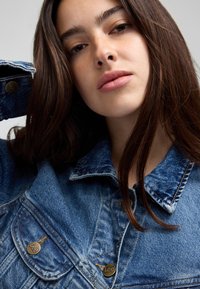 Jonge vrouw met lang bruin haar, gekleed in een blauwe spijkerjas, kijkt recht in de camera met een neutrale uitdrukking.