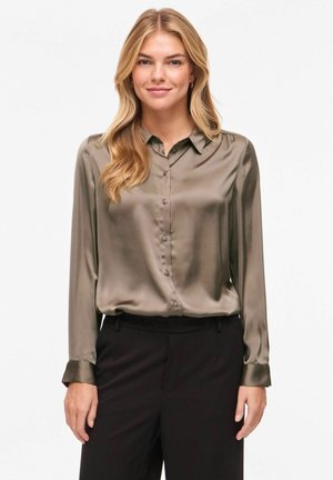 Femme blonde portant un chemisier vert olive brillant boutonné et un pantalon noir, debout devant un fond clair uni.