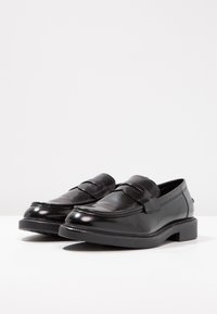 Mocassins de couro preto com acabamento polido, apresentando uma faixa proeminente e costura decorativa. Sola de borracha ligeiramente elevada.