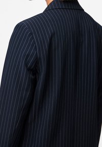 Personne portant un blazer bleu marine à fines rayures, vue de dos, mettant en avant les détails du haut du dos et des épaules.