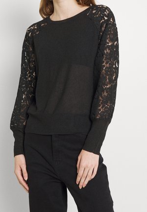 Woman wearing a black sweater with floral lace sheer sleeves and solid body, paired with black pants against a plain background.