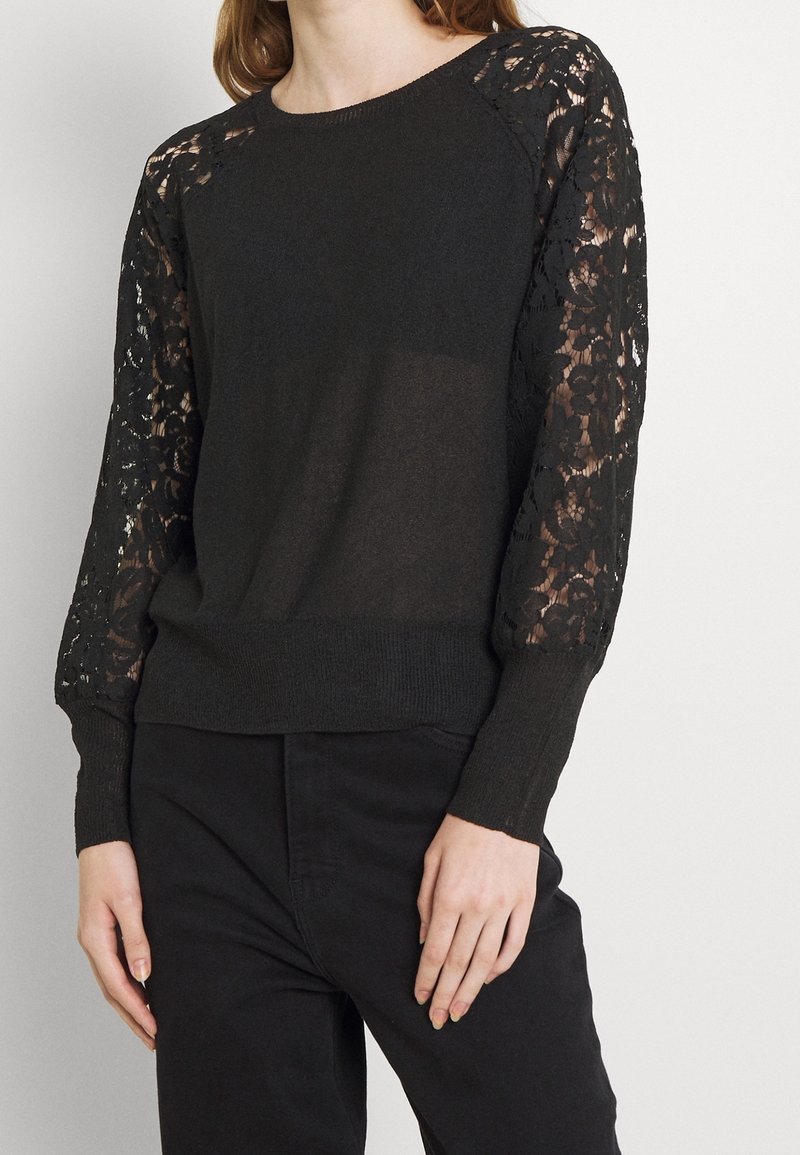 Woman wearing a black sweater with floral lace sheer sleeves and solid body, paired with black pants against a plain background.