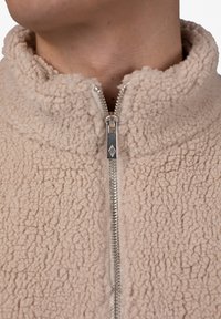 Beige Zip-Jacke aus strukturiertem Fleece-Material, mit hohem Kragen und silbernem Reißverschluss, versehen mit einem Logo am Zipper.