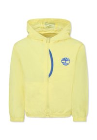 Giacca gialla con cappuccio, dotata di zip completa, polsini elasticizzati e un logo blu sul lato sinistro del petto. Tessuto leggero con una texture liscia.