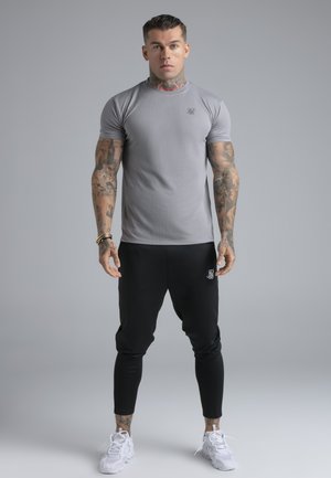 Bărbat cu tatuaje purtând un tricou gri mulat, pantaloni jogger negri și adidași albi, stând în picioare pe un fundal gri simplu.