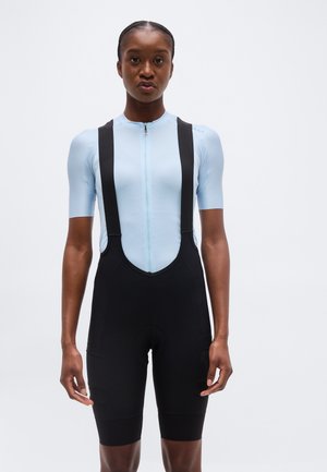 Žena oblečená v svetlomodrom cyklistickom tričku s krátkym rukávom, predným zipsom a čiernych cyklistických kraťasoch s trakmi na jednoduchom pozadí.