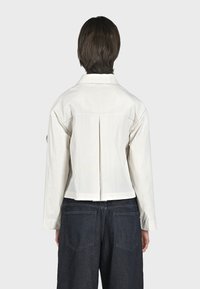 Giacca bianca cropped con apertura sul retro, colletto sartoriale e maniche lunghe. Abbinata a pantaloni in denim blu scuro, a vestibilità morbida.