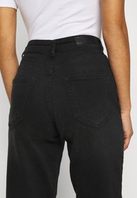 Pantalones vaqueros de denim negro con cintura alta, que presentan dos bolsillos traseros, costuras sutiles y un parche de cuero en la cinturilla.