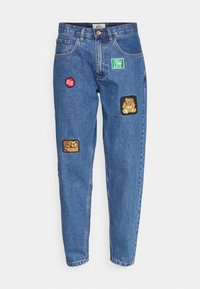 Ljusblå denimjeans med avslappnad passform, med fyra distinkta lappar: en grön fyrkant, en röd cirkel och två rektangulära lappar med mönster.