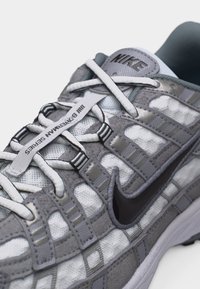 Γκρι και λευκό αθλητικό παπούτσι Nike με λεπτομέρειες από πλέγμα και σουέντ, μαύρο σήμα Swoosh και κορδόνια με το κείμενο "Bowerman Series".